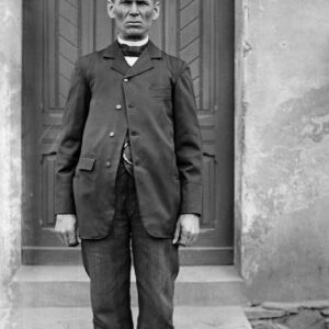 Starszy mężczyzna w szakiecie (marynarce o charakterystycznym staromodnym kroju) i sztuczkowych spodniach na tle drzwi wejściowych do domu. Oleszka (niem. Oleschka), około 1930 r.